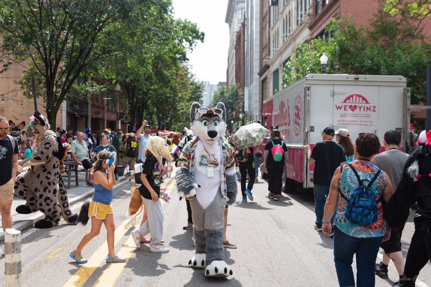 A grey wolf fursuiter in a tropical shirt and sunglasses on its brow walking down the Anthrocon Block Party, surrounded by citizens of Pittsburgh.